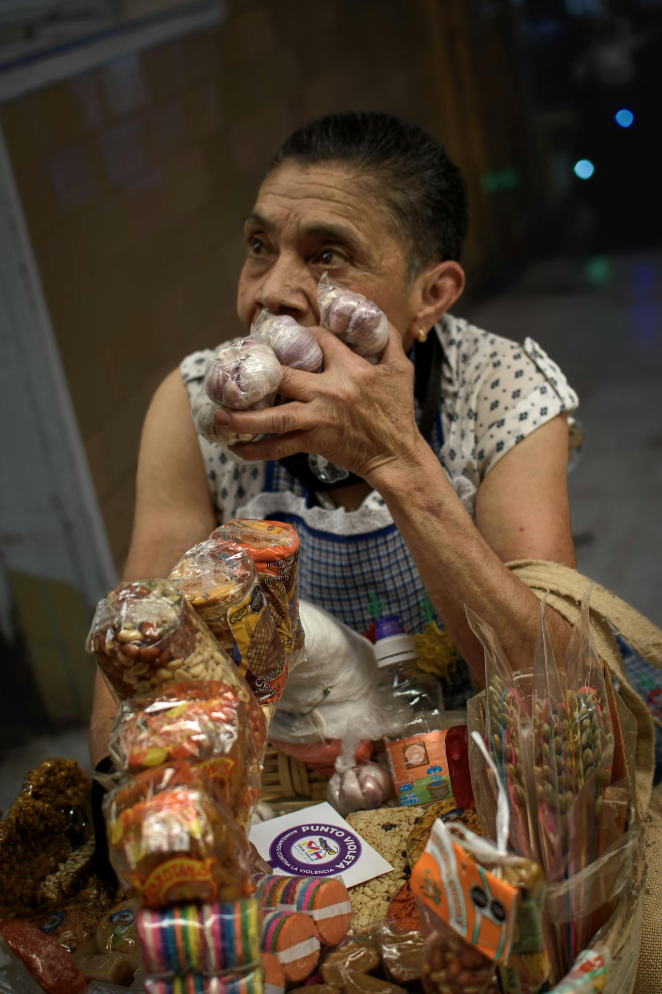 A senior woman vendor showcasing handmade items and snacks in an indoor market setting.