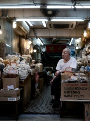 An elderly man sitting in an Asian dry goods store filled with boxes and bags at night.