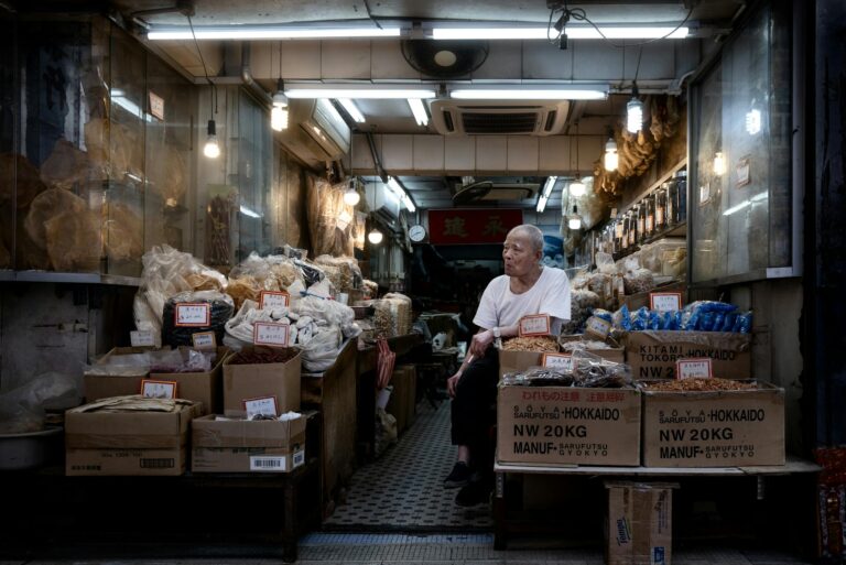 An elderly man sitting in an Asian dry goods store filled with boxes and bags at night.